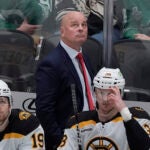 Boston Bruins head coach Jim Montgomery looks on from the bench with centers John Beecher (19) and Patrick Brown (38) during the first period of an NHL hockey game against the Dallas Stars Thursday, Nov. 14, 2024, in Dallas.