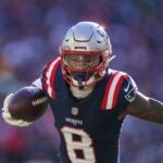 Patriots wide receiver Stefon Diggs holds a football close to his body with one arm and stiff-arming a player out of shot with the other during a game.